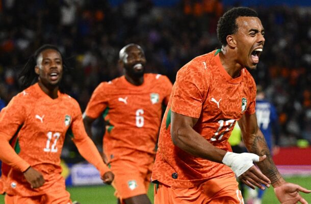 Three Senegal national team players in orange kits celebrate a goal on the soccer field, foreground player shouting with arms outstretched.
