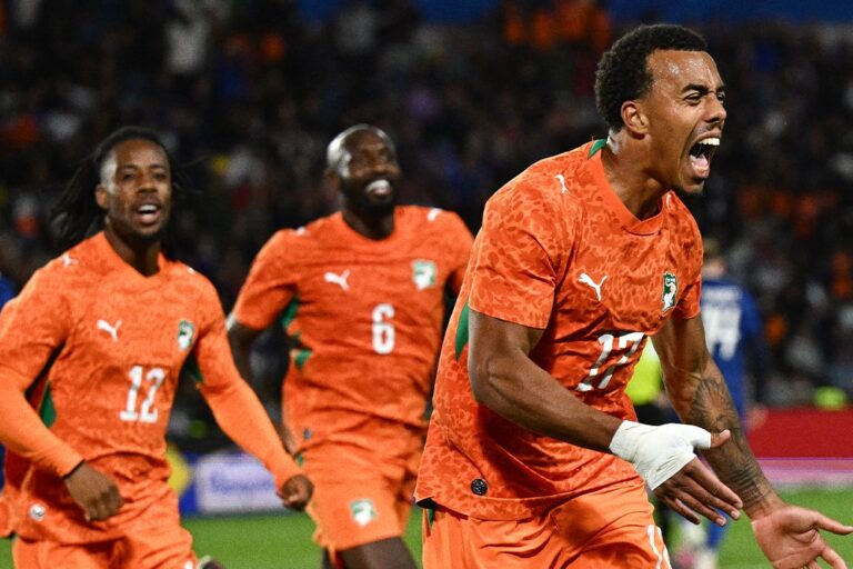 Three Senegal national team players in orange kits celebrate a goal on the soccer field, foreground player shouting with arms outstretched.