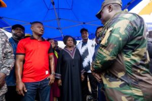 Group of civilians listening to a soldier in camouflage under a blue canopy at an outdoor event.