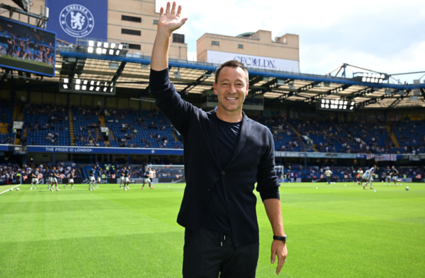 John Terry | Ghanaguardian.com Man in a dark blazer waves at the camera on a football pitch, with Chelsea FC stands and spectators in the background.