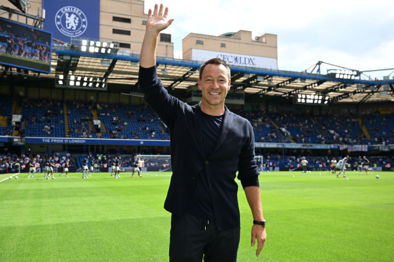 Man in a dark blazer waves at the camera on a football pitch, with Chelsea FC stands and spectators in the background.