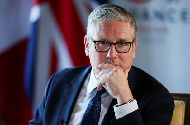 Keir Starmer | Ghanaguardian.com Man in a dark suit and glasses sits thoughtfully with his hand on his chin, at a formal event with a flag in the background.