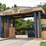 Entrance arch of Kumasi Academy with blue columns and gate, crest above, and trees beyond.