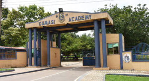 Entrance arch of Kumasi Academy with blue columns and gate, crest above, and trees beyond.