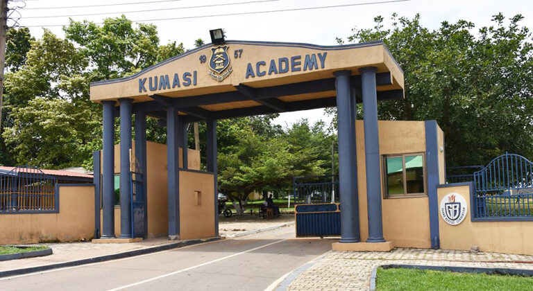 Entrance arch of Kumasi Academy with blue columns and gate, crest above, and trees beyond.