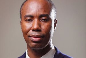 Close-up portrait of a man in a navy blazer and white shirt, looking at the camera with a calm, neutral expression.