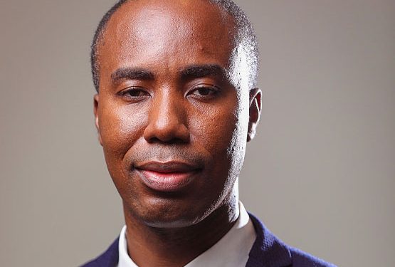 Close-up portrait of a man in a navy blazer and white shirt, looking at the camera with a calm, neutral expression.