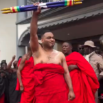 Man wrapped in a red cloth raises a multicolored staff above his head at a public ceremony, with onlookers in the background.