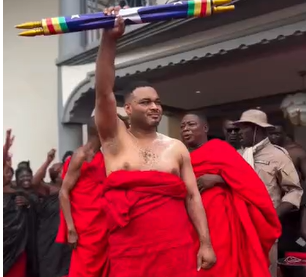 Kwadwo-Safo- | Ghanaguardian.com Man wrapped in a red cloth raises a multicolored staff above his head at a public ceremony, with onlookers in the background.