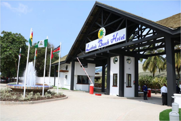 Entrance to a beach hotel with a fountain, flagpoles, and a covered driveway; sign reads 'Beach Hotel' on the front building.