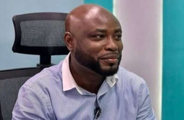 Smiling Black man with a trimmed beard sits in an office chair wearing a light blue striped shirt, microphone clipped to his shirt.