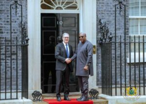 Two men in formal attire shake hands on a red carpet outside a black door marked with the number 10, in front of a stately brick building.