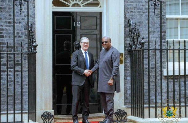 Two men in formal attire shake hands on a red carpet outside a black door marked with the number 10, in front of a stately brick building.