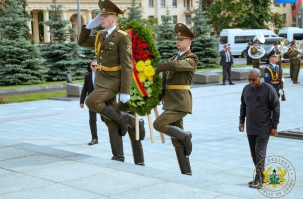 Mahama Minsk | Ghanaguardian.com Two soldiers in dress uniforms march in formation, carrying a large red-and-yellow floral wreath during a ceremonial event.