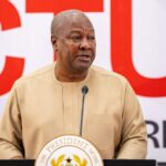 African man in a beige traditional shirt speaking at a podium with a microphone in front of a bold red-and-white backdrop and presidential seal