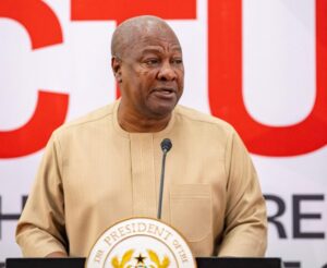 African man in a beige traditional shirt speaking at a podium with a microphone in front of a bold red-and-white backdrop and presidential seal