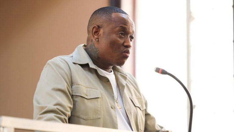 Man at a podium speaking into a microphone, wearing a beige jacket and white shirt.