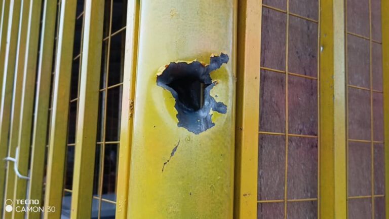 Close-up of a yellow metal gate with a large torn hole and visible cracks in the panel.