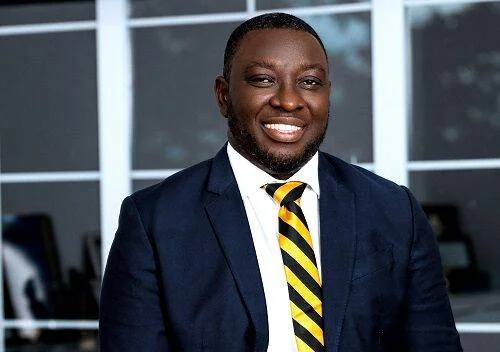 Smiling man in a navy suit and yellow-black striped tie standing in a modern office setting