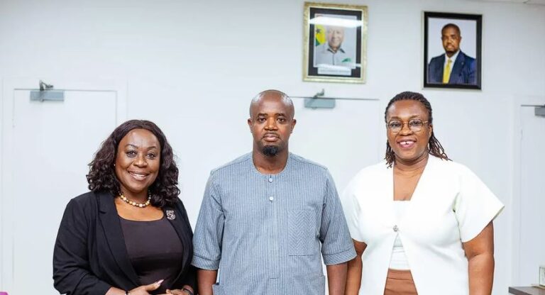 Three professionals posing in an office: man center in a light blue tunic, women on either side in dark and white outfits; framed portraits on the wall behind.