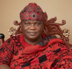 Portrait of a man in a red and black patterned traditional outfit and matching cap, seated on a carved wooden chair with floral motifs, wearing a silver wristwatch on his left wrist.