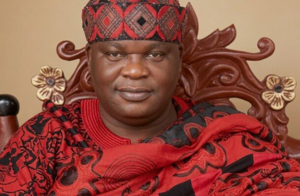 Portrait of a man in a red and black patterned traditional outfit and matching cap, seated on a carved wooden chair with floral motifs, wearing a silver wristwatch on his left wrist.
