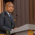 Man in a dark suit and orange pocket square speaks at a podium with a microphone, ministry seal visible on the lectern.
