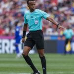 Soccer referee in turquoise shirt and black shorts signaling on a green pitch as fans watch in the stands.