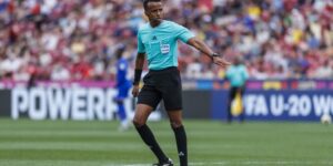 Soccer referee in turquoise shirt and black shorts signaling on a green pitch as fans watch in the stands.