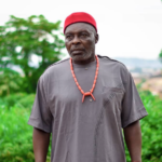 Older man in a red cap and gray traditional tunic with a coral necklace, outdoors with greenery in the background.