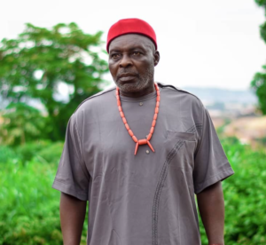 Older man in a red cap and gray traditional tunic with a coral necklace, outdoors with greenery in the background.