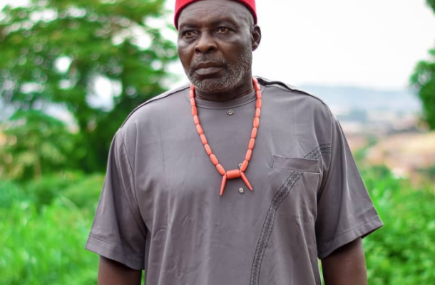 Patrick Okoye | Ghanaguardian.com Older man in a red cap and gray traditional tunic with a coral necklace, outdoors with greenery in the background.