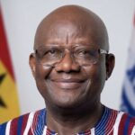 Smiling African man with glasses, wearing a striped shirt, with national flags in the background.