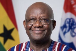 Smiling African man with glasses, wearing a striped shirt, with national flags in the background.