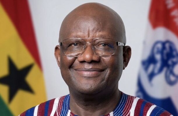 Smiling African man with glasses, wearing a striped shirt, with national flags in the background.