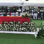 Outdoor funeral or memorial ceremony under a white canopy; a red casket is centerpiece, surrounded by white floral arrangements on a green lawn, with attendees seated in the background.