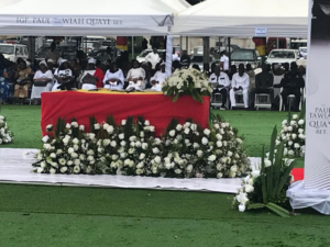 Outdoor funeral or memorial ceremony under a white canopy; a red casket is centerpiece, surrounded by white floral arrangements on a green lawn, with attendees seated in the background.