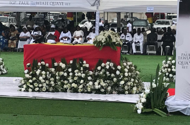 Paul Tawiah Quaye | Ghanaguardian.com Outdoor funeral or memorial ceremony under a white canopy; a red casket is centerpiece, surrounded by white floral arrangements on a green lawn, with attendees seated in the background.
