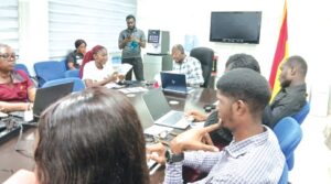 People gathered around a conference table with laptops, having a team meeting in a bright room with a flag in the corner.