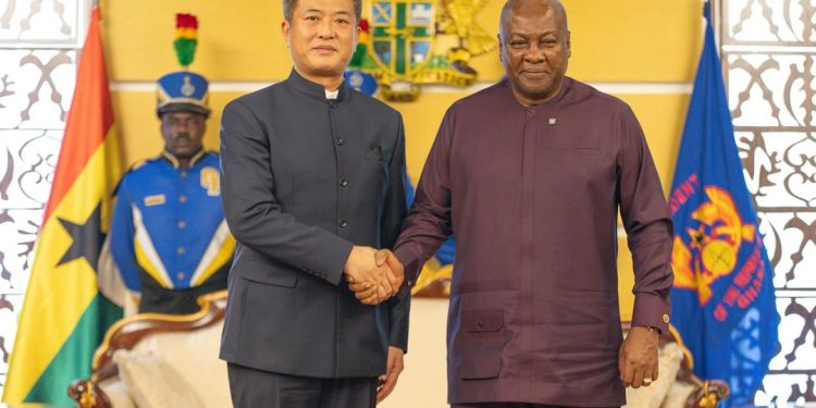 Two men in formal attire shake hands in a ceremonial room, with national flags and a uniformed guard in the background.