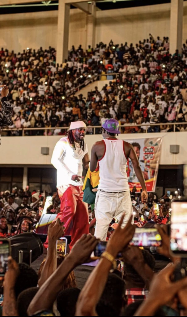 Two performers on a lit stage facing each other as a large crowd watches from stands, many people holding up phones to record.