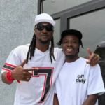 Two men posing outside a building; left man wears a white cap, sunglasses, and a white jersey with a large number 7, right man wears a white T‑shirt that says 'off duty'.