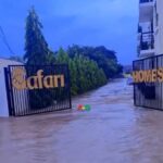 Flooded driveway at gated entrance with gates reading 'Safari' and 'Homes', water covering the path and trees lining the sides.