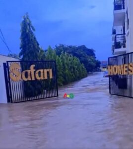 Flooded driveway at gated entrance with gates reading 'Safari' and 'Homes', water covering the path and trees lining the sides.