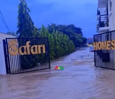 Flooded driveway at gated entrance with gates reading 'Safari' and 'Homes', water covering the path and trees lining the sides.