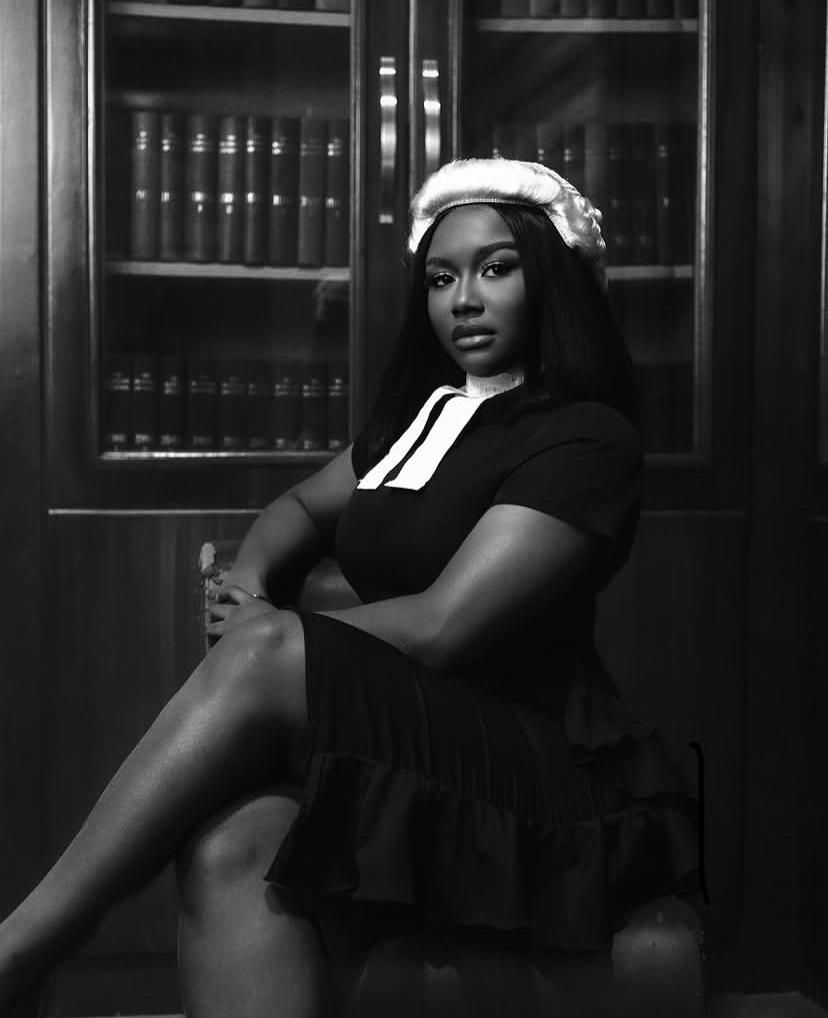 Black-and-white portrait of a woman in a barrister wig and black dress seated in front of a bookshelf of legal volumes in a law library.