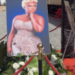 Portrait of a woman in a sparkling strapless dress on a stand, surrounded by white flowers and red rope barriers at an outdoor event