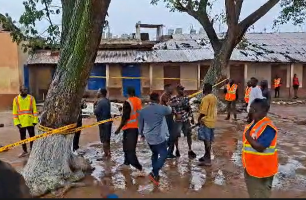 Screenshot-2026-06-06-134337 | Ghanaguardian.com Group of people in orange safety vests gathered in a muddy, taped-off area near a large tree and row of huts in a yard.