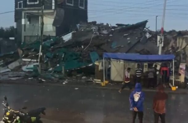 Rubble and debris of a collapsed building on a street, firefighters on the left, and a blue canopy with bystanders nearby under overcast sky.