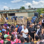 A large community gathering outdoors as a man in a white shirt speaks to a crowd, with wooden planks and vehicles in the background.